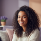 Smiling woman with dark curly hair and warm brown skin in a candid portrait headshot, against a blurred background with soft 